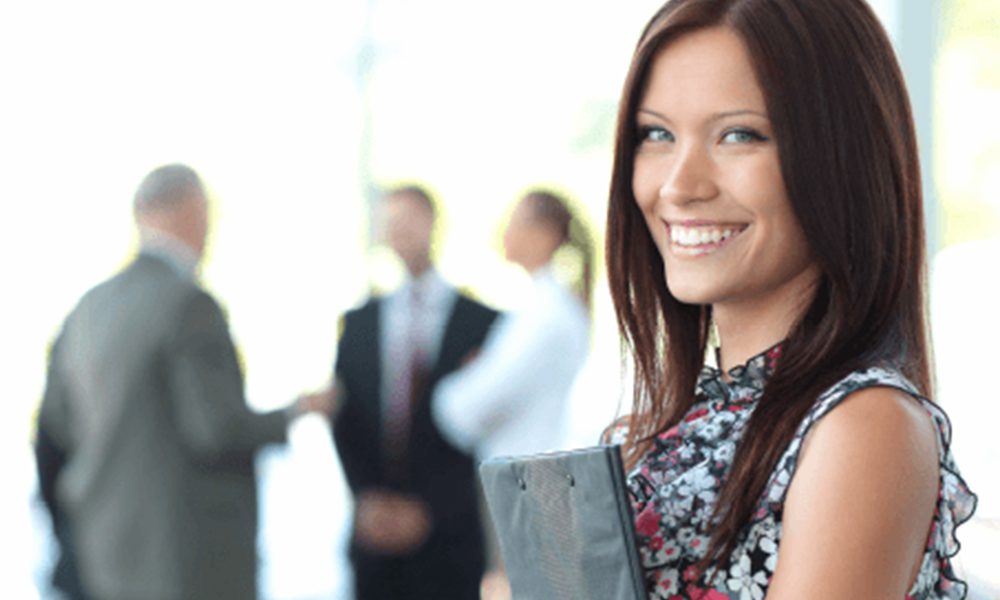 Employee Benefits Assistant Account Manager - Professional Woman Smiling and Looking at Camera with a Portfolio in her Hands and Professionals Talking in Background