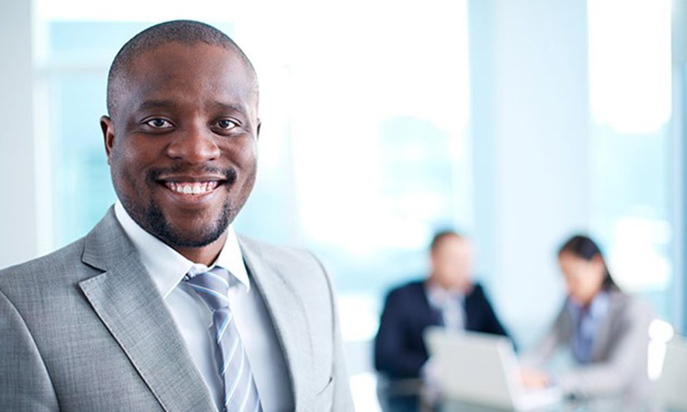 Employee Benefits Consultant - Professional Man Smiling and Starring at the Camera as People Sit in a Conference Room in an Office Blurred Behind Him