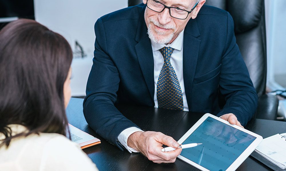 Private Client Sales Representative - Older Professionally Dressed Man Pointing out Something on a Tablet to a Woman While They Sit in the Office