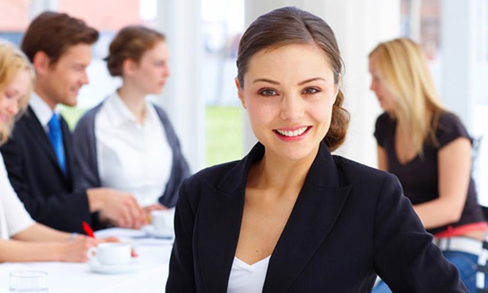 Property & Casualty Risk Management Consultant - Professional Woman Smiling and Starring at the Camera While Her Coworkers Talk in the Background at an Office Table